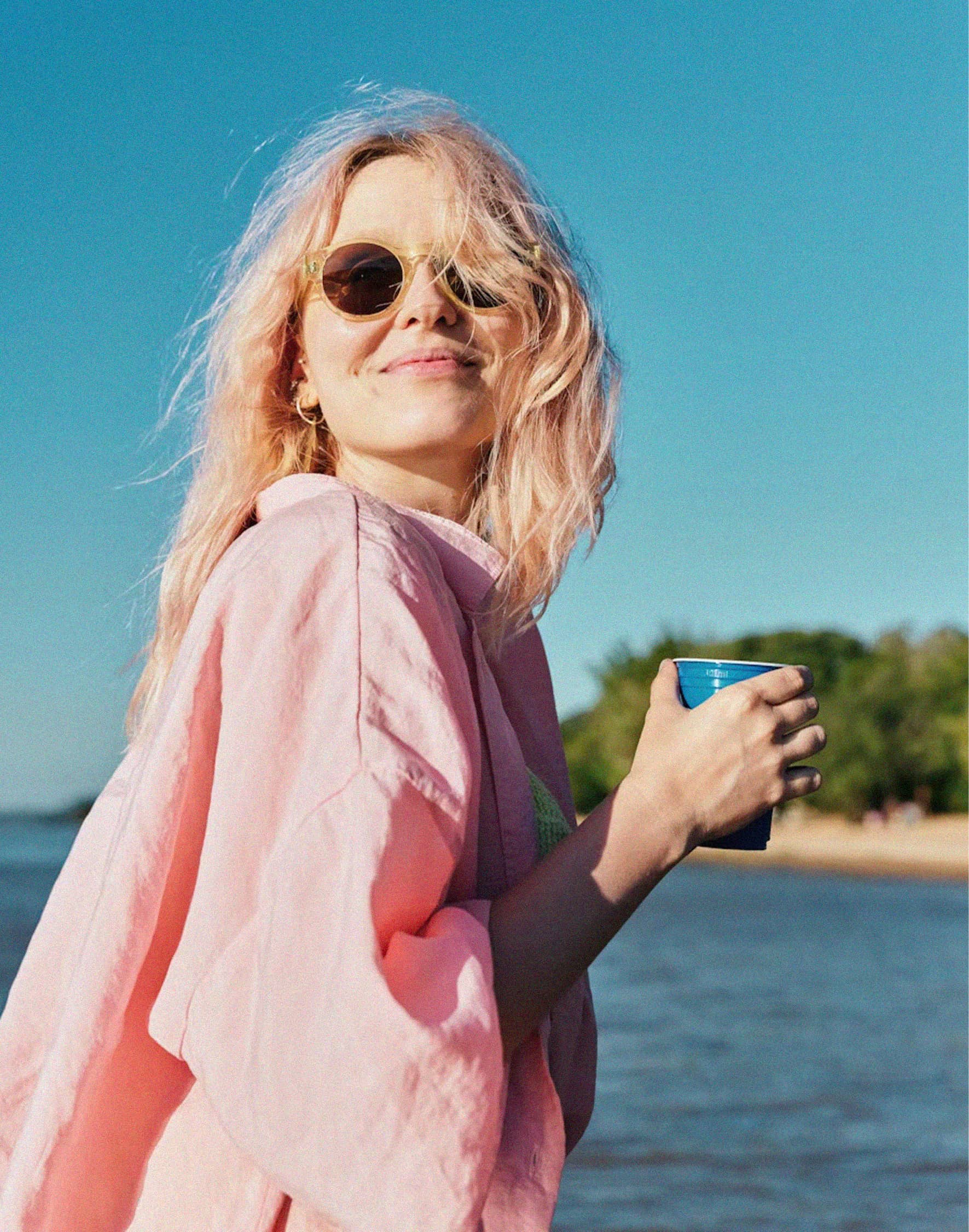 A woman wearing sunglasses and an outdoor jacket, holding a mug.