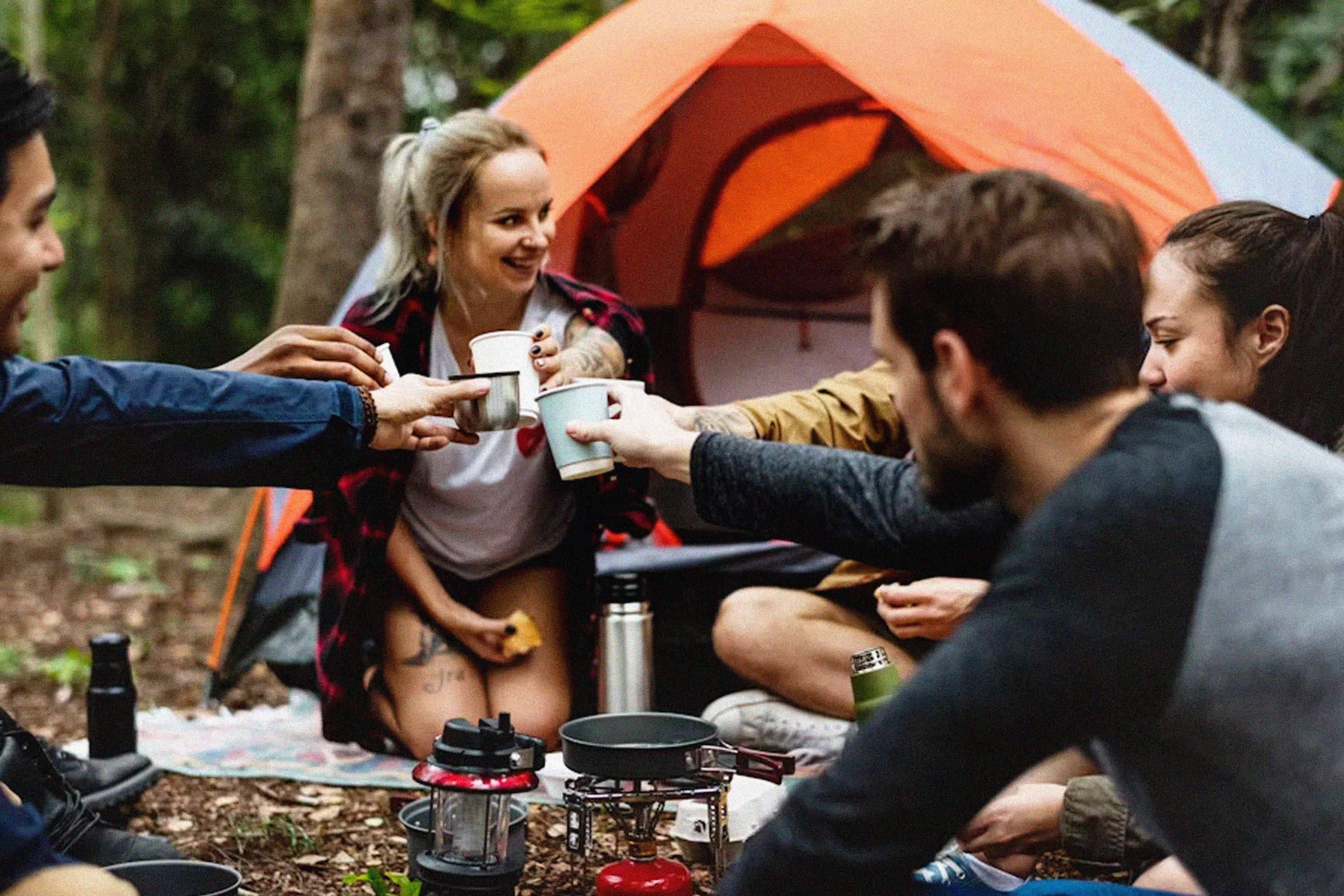 Friends raising camping mugs around an outdoor table beside a tent.